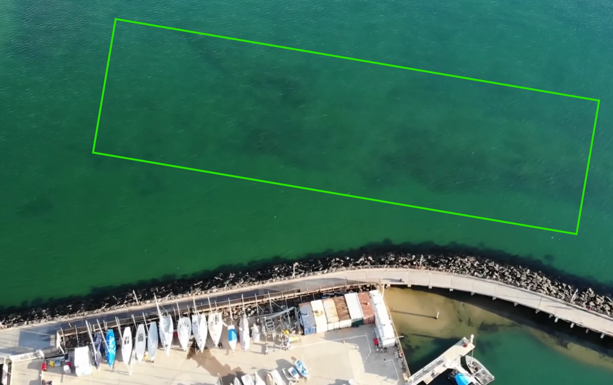 Aerial view of the Brighton breakwall showing the rock wall, the elevated concrete pier section and a marked zone of open water with darker weed patches